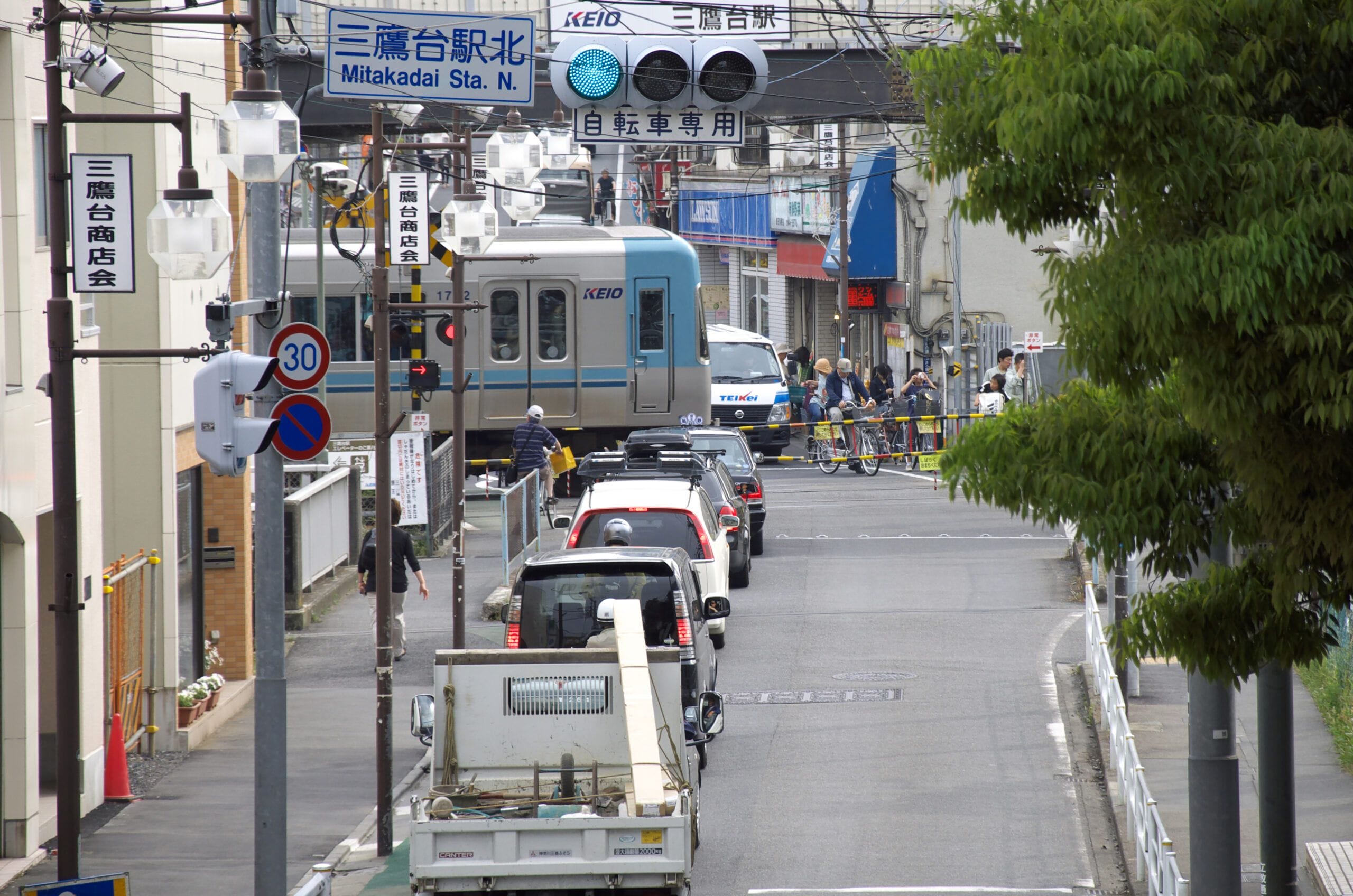 日本の街路。踏切で電車が通過するのを待って車が停車し、歩行者が待機している様子が見られ、通りや駅の様々な標識が確認できる。.