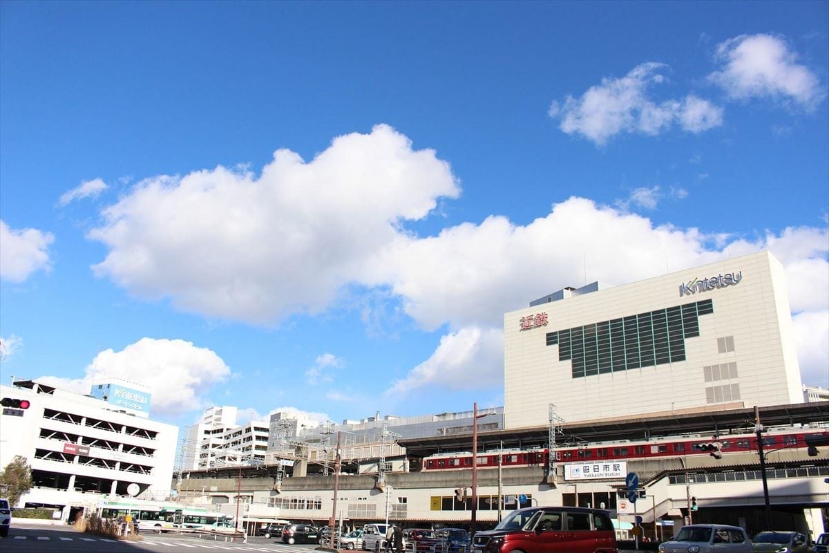 「近鉄」の看板が掲げられた大きな駅舎、駐車場、そしてその前には数台の車やバスが停まっており、空は青く、所々雲がかかっている。.