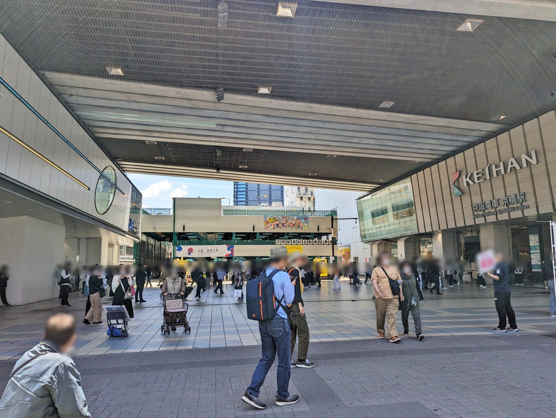 近代的なビル群に囲まれた屋外の広場では、人々が歩き回ったり立ち止まったりしている。右側には京阪の看板が見え、頭上には屋根付きの構造物がそびえ立っている。.
