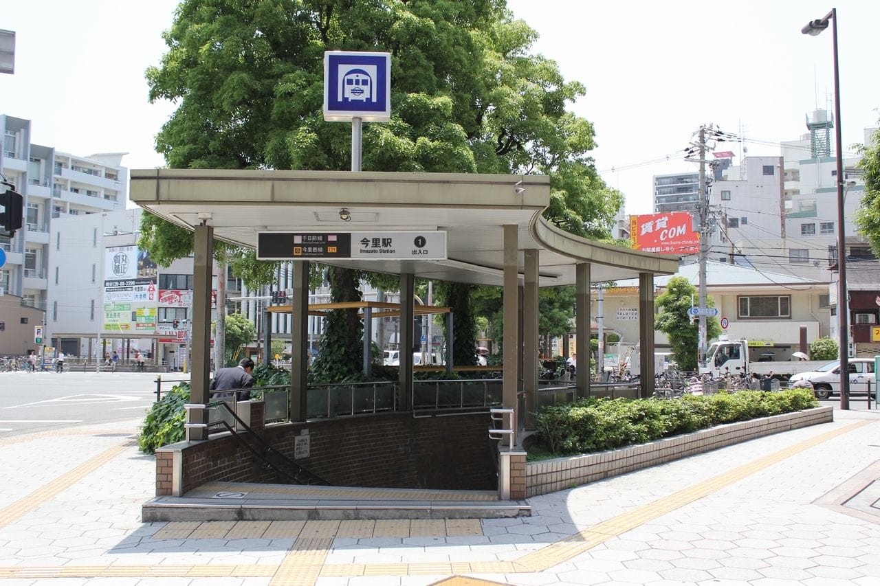 晴れた日の、頭上に看板が掲げられた地下鉄駅の入り口。周囲は緑に囲まれ、背景には街のビル群が映っている。.