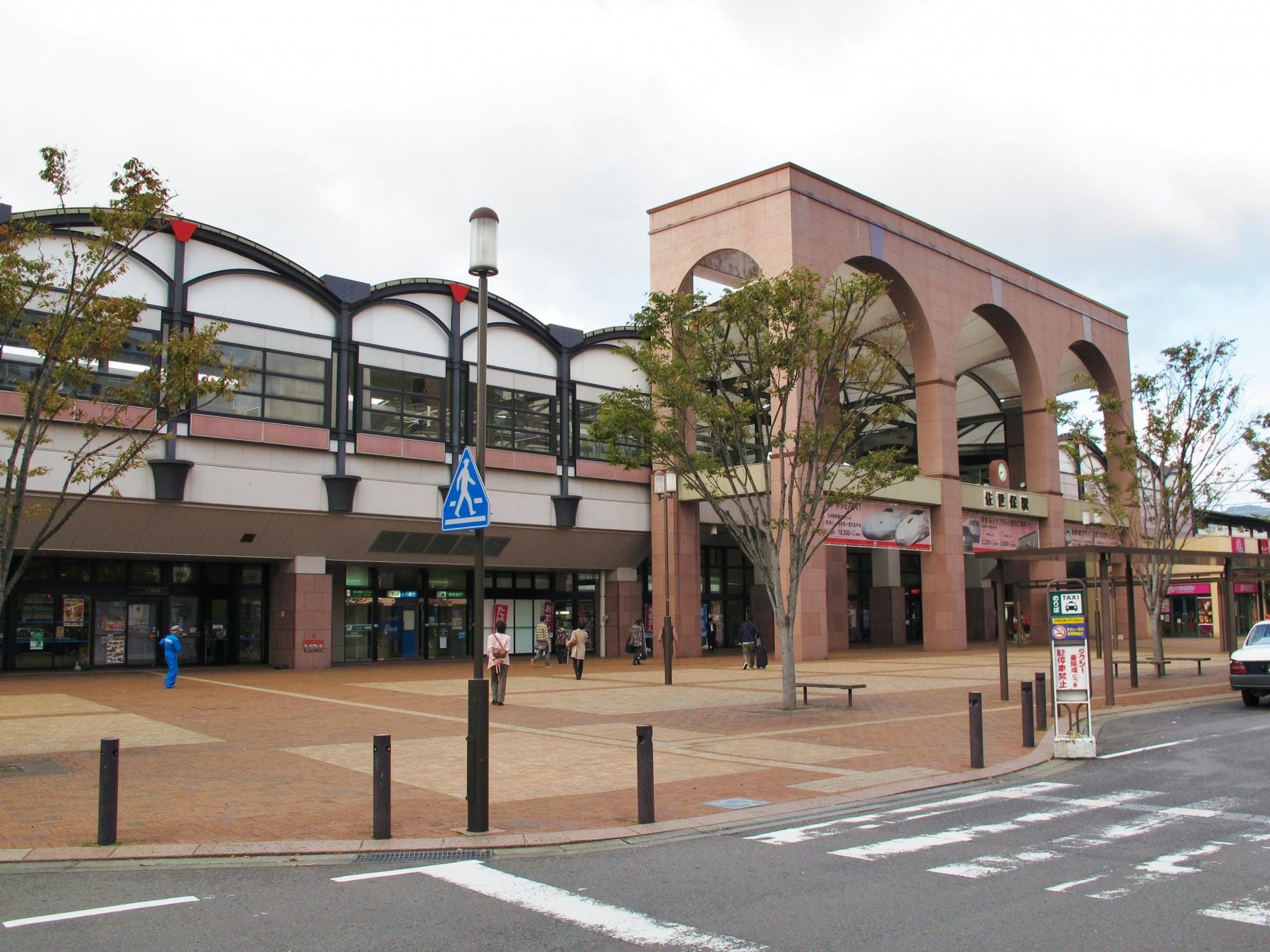 アーチ型の入り口を備えたモダンな駅舎。その前には歩行者専用エリアが広がり、木々が立ち並び、入り口付近を数人の人々が歩いている。.