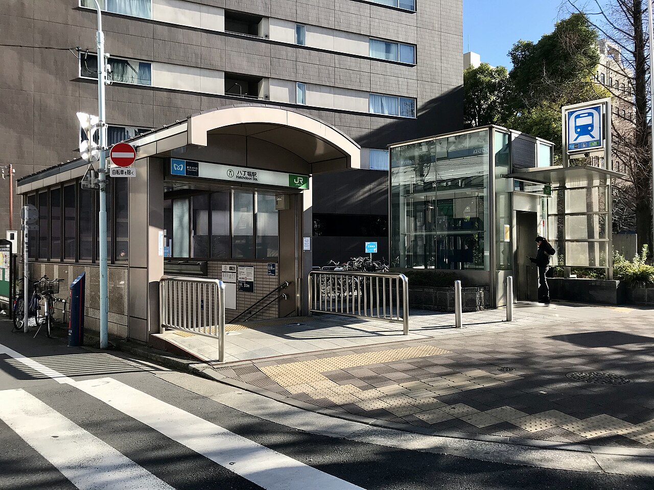 ガラス扉、手すり、そして列車のアイコンが描かれた看板がある地下鉄駅の入り口。背景には建物と木々が写っている。.