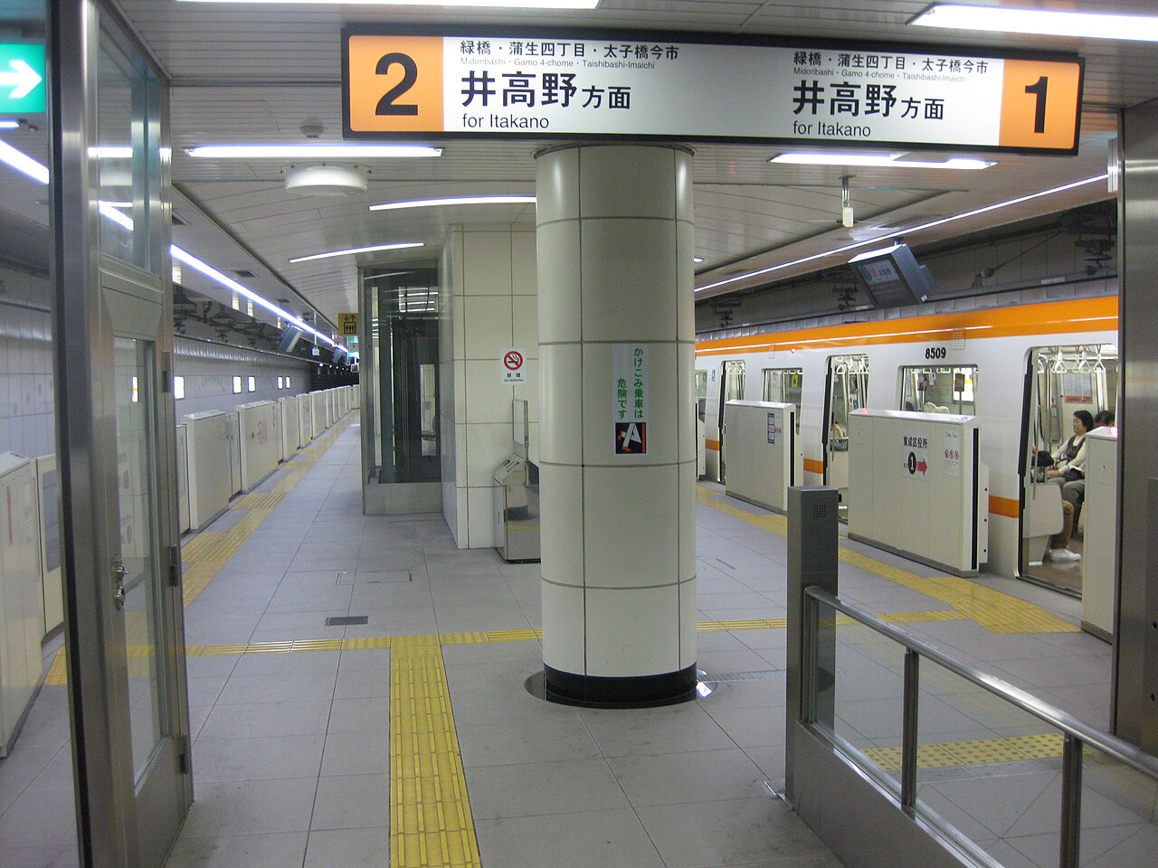 東京の地下鉄のホームには、黄色い点字ブロックが敷かれ、板野方面行きの1番線と2番線を示す案内表示があり、ホームには電車が停車している。駅は清潔で、照明も明るい。.